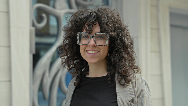 Woman wearing oversized tortoiseshell glasses smiles at camera showing teeth while standing by decorative metal sculpture and stone facade on street; confidence warmth.