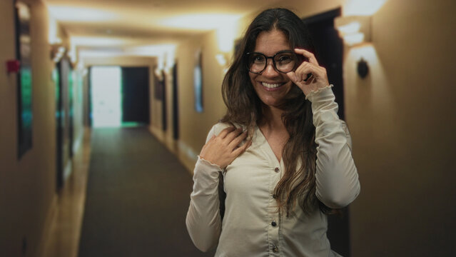 Woman adjusts glasses and smiles in a hotel building corridor lit by warm lamps; confidence friendliness approachability.