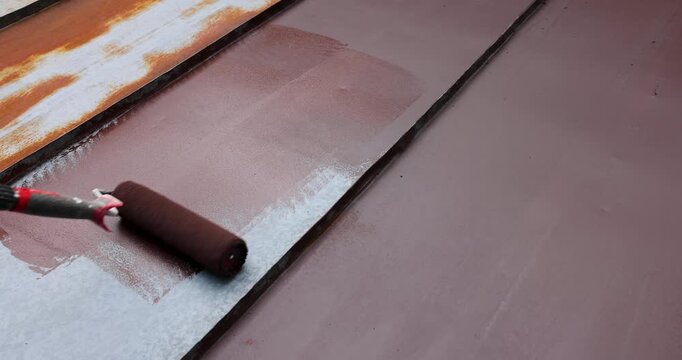 painting tin metal roof with roller and brown paint. rusty house roofing restoration