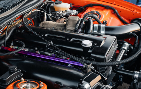 Engine bay an orange 1990s sports car with inline six cylinder engine. Close-up of a modified motor with black cylinder head and intake piping under the hood.