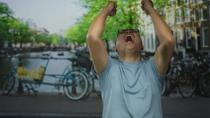 Obraz premium Man cheering with raised fists on a street beside an amsterdam canal, wearing glasses and a blue tee shirt; victory joy.