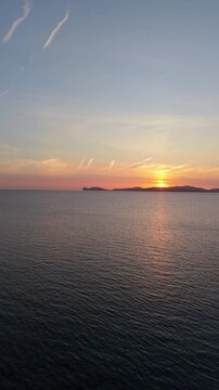 Vertical aerial pull-back view of Capo Caccia at sunset revealing dramatic cliffs and Mediterranean sea in warm golden hour light.