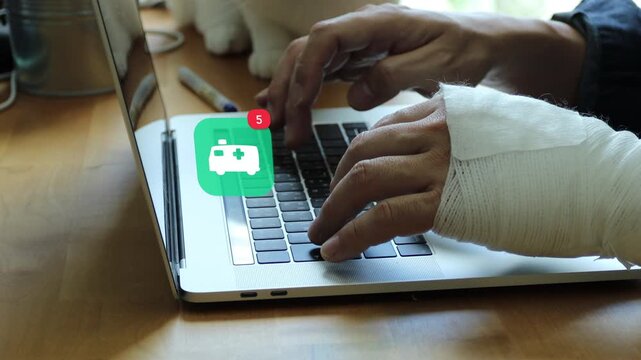 Close up of an injured East Asian man with a bandaged arm typing on a laptop, showing a 3D tracked emergency medical ambulance notification pop up and red counter.