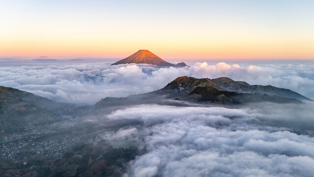 Aerial view of a majestic volcano rising above a sea of clouds during the golden hour with rugged mountain slopes visible below in Indonesia.