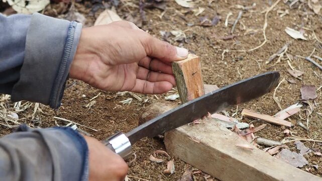 Close-up of hands using a machete to carve and shape a piece of wood, demonstrating traditional woodworking outdoors.