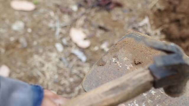 Close-up of a person using a hammer to drive a wooden wedge into the handle of an old, rusty garden hoe for repair and maintenance.