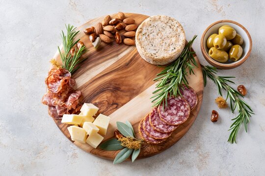 Charcuterie ingredients placed around empty serving board for cheese and meat platter preparation. Various ingredients are arranged around an empty serving board. Finer food snacks