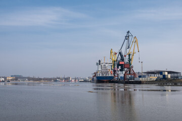 Fototapeta premium Industrial harbor features a cargo ship docked next to large cranes and warehouses, with a clear blue sky and calm water reflecting the scene