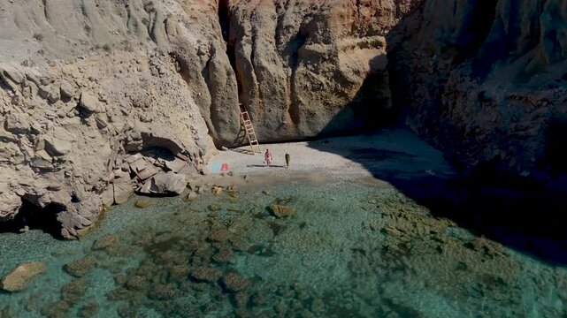 Discover the stunning coastline of Milos, Greece, featuring crystal-clear waters and dramatic rock formations, with a couple of men and woman walks at the Tsigrado Beach