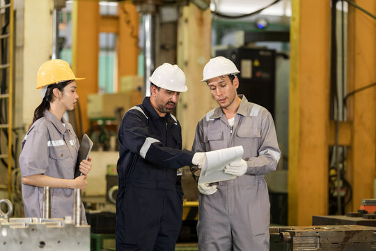 Three adult engineers review printed report beside tooling area, discussing process detail, production status and planning steps during collaborative factory meeting in workshop.