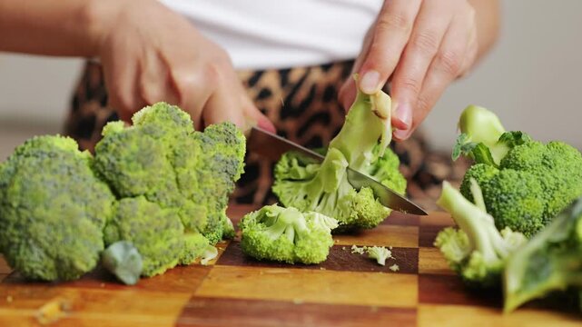A person holds a knife to cut broccoli on a wooden cutting board. The kitchen has natural light and a simple setting. Fresh vegetables are arranged neatly