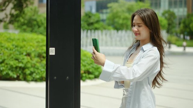 Smiling young Asian business woman scanning a QR code on a modern outdoor interactive digital touch screen kiosk using a smartphone, experiencing seamless mobile technology and smart city connectivity