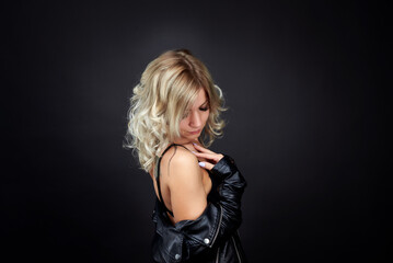 Fototapeta premium A blonde woman in a black leather jacket and blue jeans is standing in a dark studio. She is wearing a black bra and has her beautiful breasts exposed.