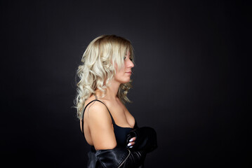 Naklejka premium A blonde woman in a black leather jacket and blue jeans is standing in a dark studio. She is wearing a black bra and has her beautiful breasts exposed.