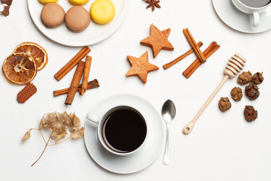 Una taza de cafe negro junto a un plato con macarons, unas ramas de canela y unas estrellas de madera decorativas sobre un fondo blanco. Vista superior y d ecerca