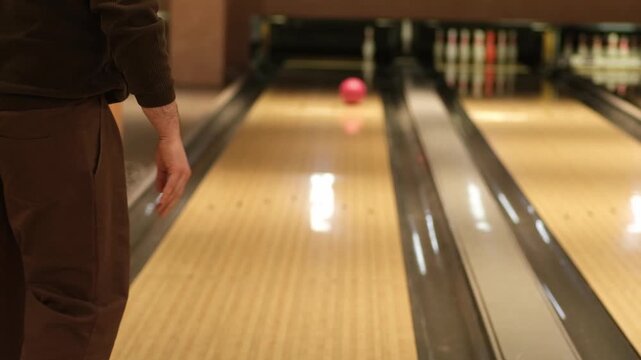 Man dressed in brown prepares to roll a pink bowling ball down the lane in a bowling alley, with bowling pins in the background