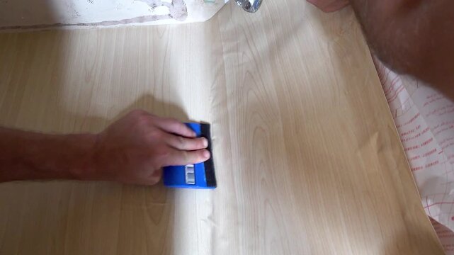 Close up view of a person using a squeegee to apply self-adhesive vinyl film onto a cabinet surface under a sink during home renovation.