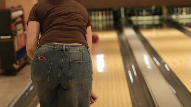 Woman in casual clothing positions herself to bowl with a pink ball in a bowling alley, showcasing the lane and bowling pins in the background