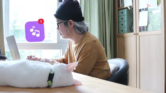 Side view of a mature Asian man using a laptop at home office next to his cat, displaying a 3D tracked musical note audio melody streaming notification pop up and red counter.