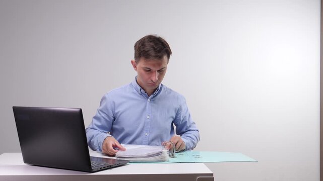 Man reviewing financial paperwork in a binder while using a laptop to process data and handle administration tasks in a modern office setup