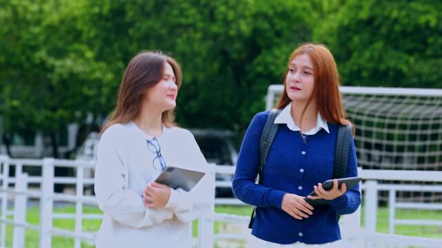 Happy student walking with classmate friend on green school campus for education, talking while holding digital tablet device cheerfully near outdoor sport field