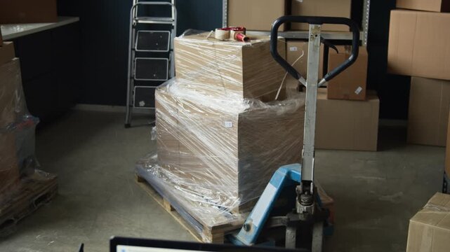 Establishing shot of laptop displaying logistics software next to safety helmet on desk, camera panning to reveal organized warehouse with boxes. Clip is ideal for supply chain and e-commerce