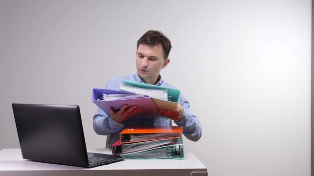 Man struggling with an overwhelming amount of paperwork and documents, holding a stack of binders while trying to work at a laptop, illustrating stress, busy tasks, and office challenges