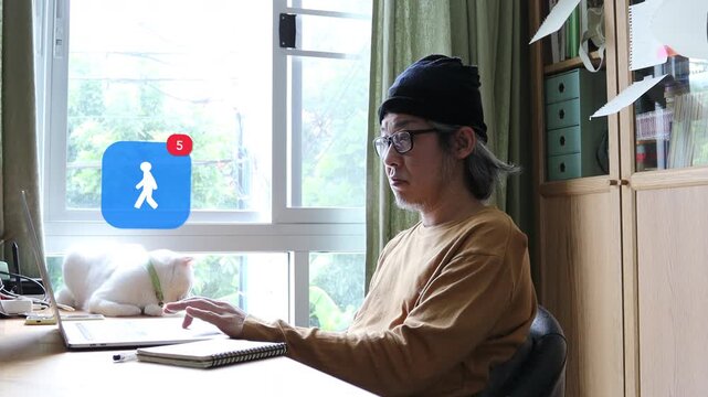 Mature Asian man with white hair using laptop at home office next to his cat, with a 3D tracked pedestrian walking silhouette fitness tracker notification pop up and red counter.