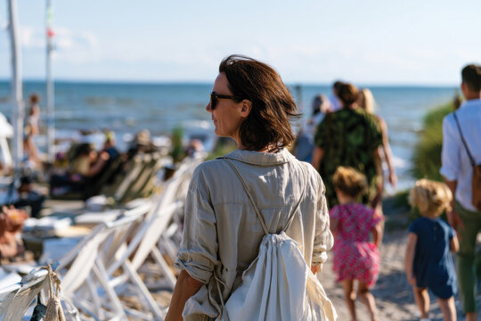 A serene image of a woman strolling along the beach, enjoying the sun, with children and other beachgoers creating a vibrant seaside atmosphere, capturing the essence of leisure.