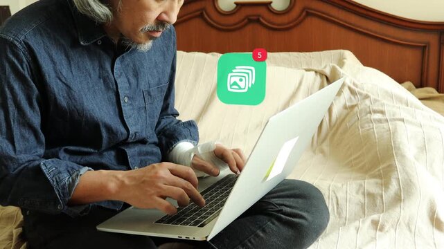 Hurt hand mature Asian man with grey beard using laptop in bedroom, showing a 3D tracked digital picture stack photo gallery notification pop up and red counter.