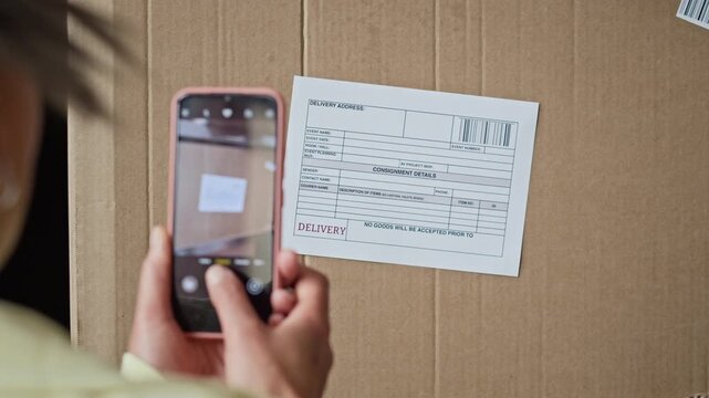 Over the shoulder view of young worker using smartphone to photograph delivery label on cardboard box in warehouse. Footage represents mobile app integration in e-commerce logistics