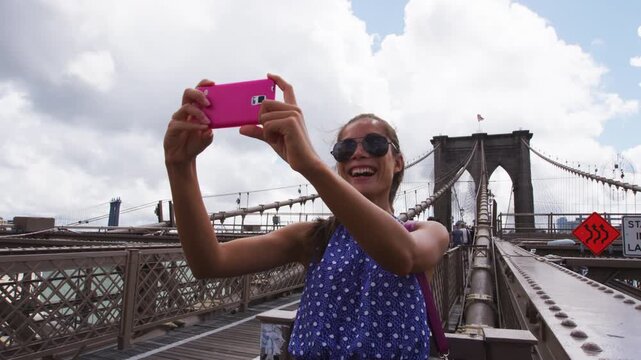 Happy selfie tourist woman taking fun self-portrait picture with smart phone app on Brooklyn Brige, New York City, Manhattan, USA. Asian woman posing with smartphone for social media.