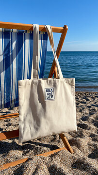 Summer beach lifestyle: Cream canvas tote bag hanging on a striped wooden beach chair against clear blue ocean.