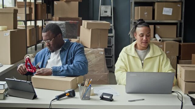 Stab shot of African American young man taping box and scanning barcode while multiethnic woman working on laptop in warehouse. Clip perfect for e-commerce and retail logistics marketing