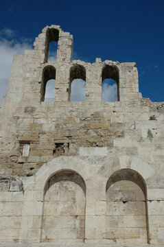The historic ruined stone facade and large arches of the Odeon of Herodes Atticus stand under a bright blue sky at the Acropolis in Athens Greece.