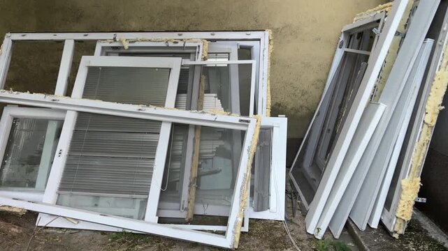 close up view of disassembled plastic windows stacked outside after being removed from a building with white frames glass window panes built in blinds and remaining yellow polyurethane foam