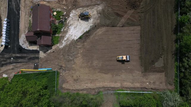 Aerial view shows a roller machine compacting and leveling the ground at a construction site near homes. Workers are preparing the area for building activities
