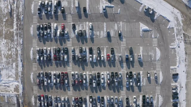 High overhead drone video slowly moving down toward a large winter parking area with rows of parked cars, snowbanks, empty spaces, pedestrian crossings and long shadows.