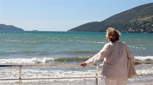 Senior woman standing by a railing and looking at waves on a windy sea with coastal mountains in the background. Mature travel and seaside lifestyle scene.