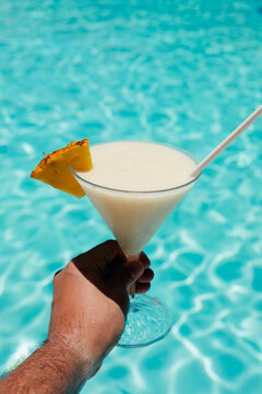 POV of a hand holding a pineapple cocktail by a glittering pool