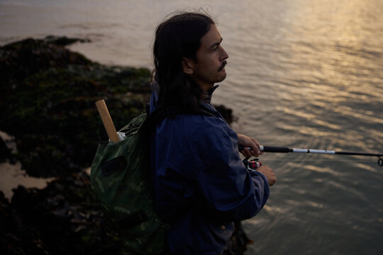 Fisherman stands on rocks by the water during sunset