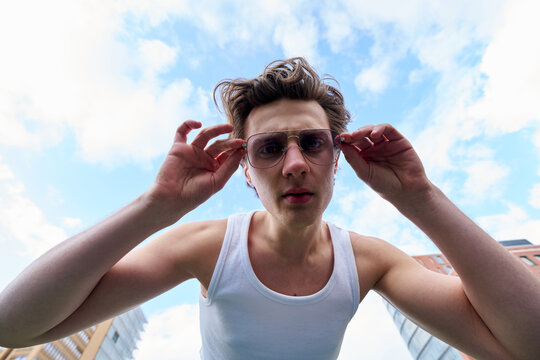 Young Man Adjusts Sunglasses Under Bright Sky in Urban Area