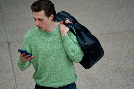 Man Looks at Phone While Carrying Bag in a Public Space