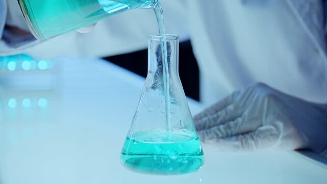 Close up shot of hands of unrecognizable chemist in sterile gloves and lab coat pouring blue liquid from glass beaker into Erlenmeyer flask on illuminated laboratory table