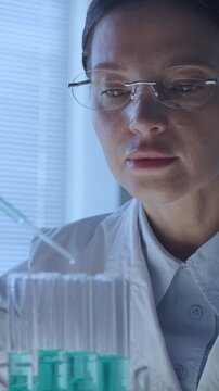 Focused female chemist in eyeglasses and lab coat using pipette to add blue liquid to row of test tubes during biotechnology research in laboratory. Vertical close-up shot