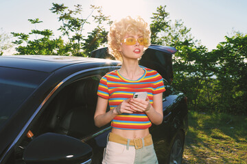 Obraz premium Young woman with curly blonde hair stands beside a sleek car in bright sunlight, wearing a colorful striped crop top and light jeans, enjoying a carefree outdoor moment.