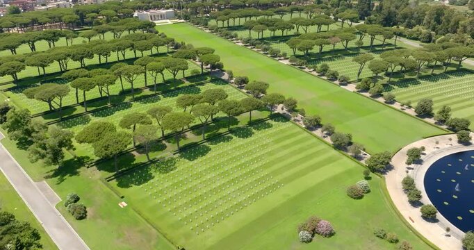 Aerial view of a military cemetery, featuring meticulously manicured green lawns with distinct striped patterns, lined by parallel rows of tall umbrella pine trees. It's located in Nettuno, Italy.