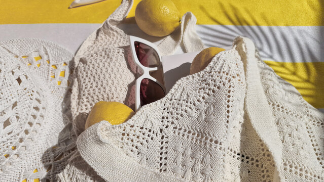 Summer vacation, summertime, travel, tourism concept. White crochet bag with fresh lemons and sunglasses on white and yellow striped beach towel. 