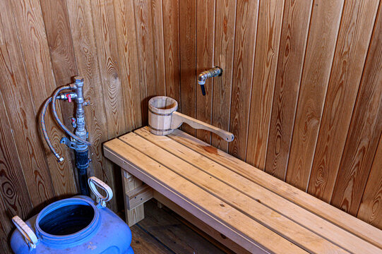 Wood-paneled sauna washroom features a wooden bench, a birch bucket with a handle, a blue water tank, and metal plumbing with a tap