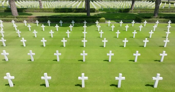 Aerial view showing endless rows of symmetrical white marble crosses standing perfectly aligned on a pristine green lawn at a historic military cemetery. Many cypress trees line the tombs.